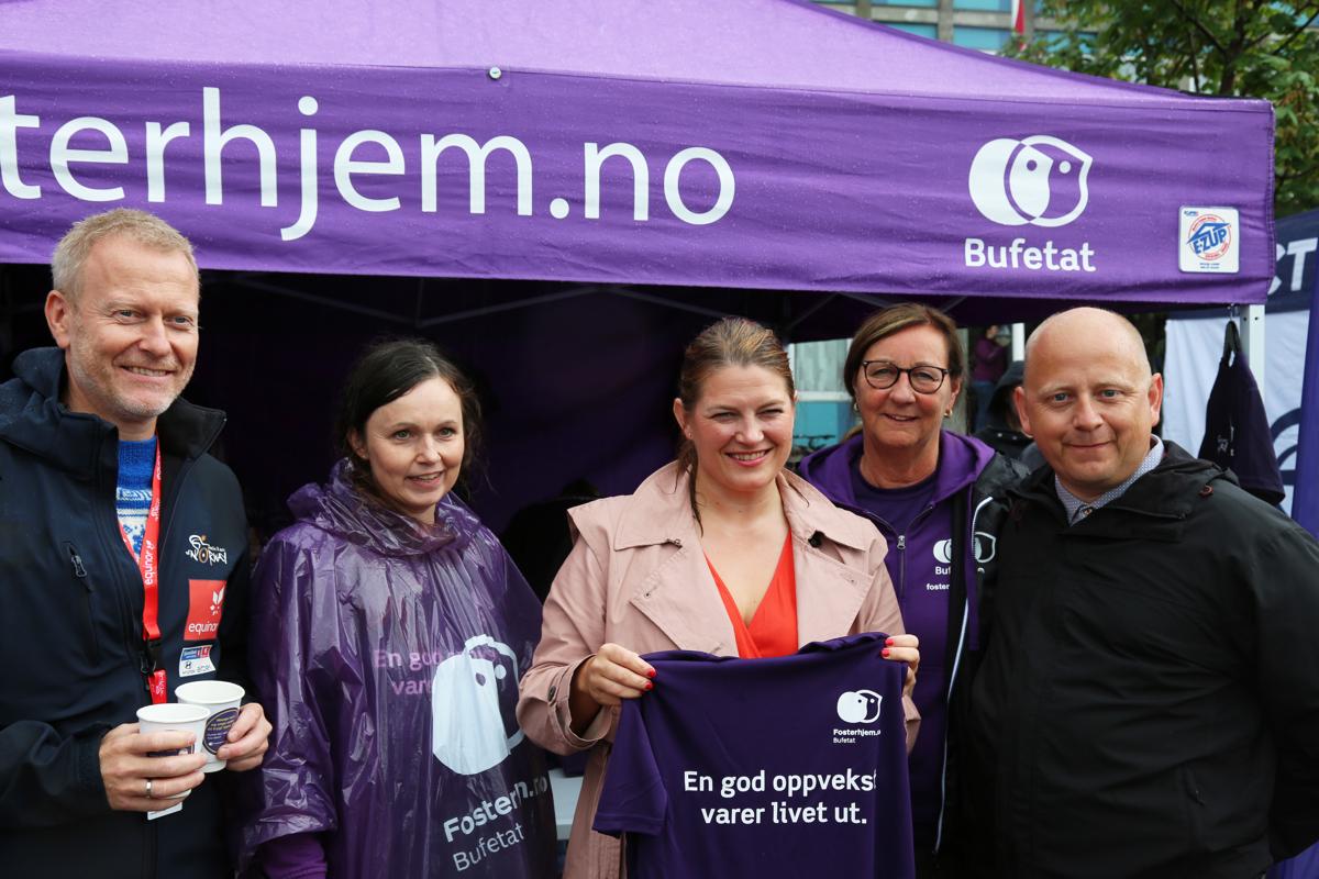 Fem mennesker foran et lilla telt med teksten fosterhjem.no på.