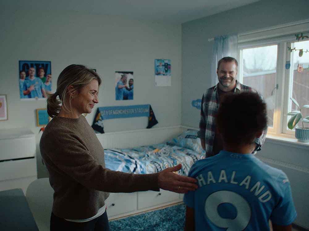 En kvinne og en mann tar imot ei jente i fotballdrakt, på et soverom