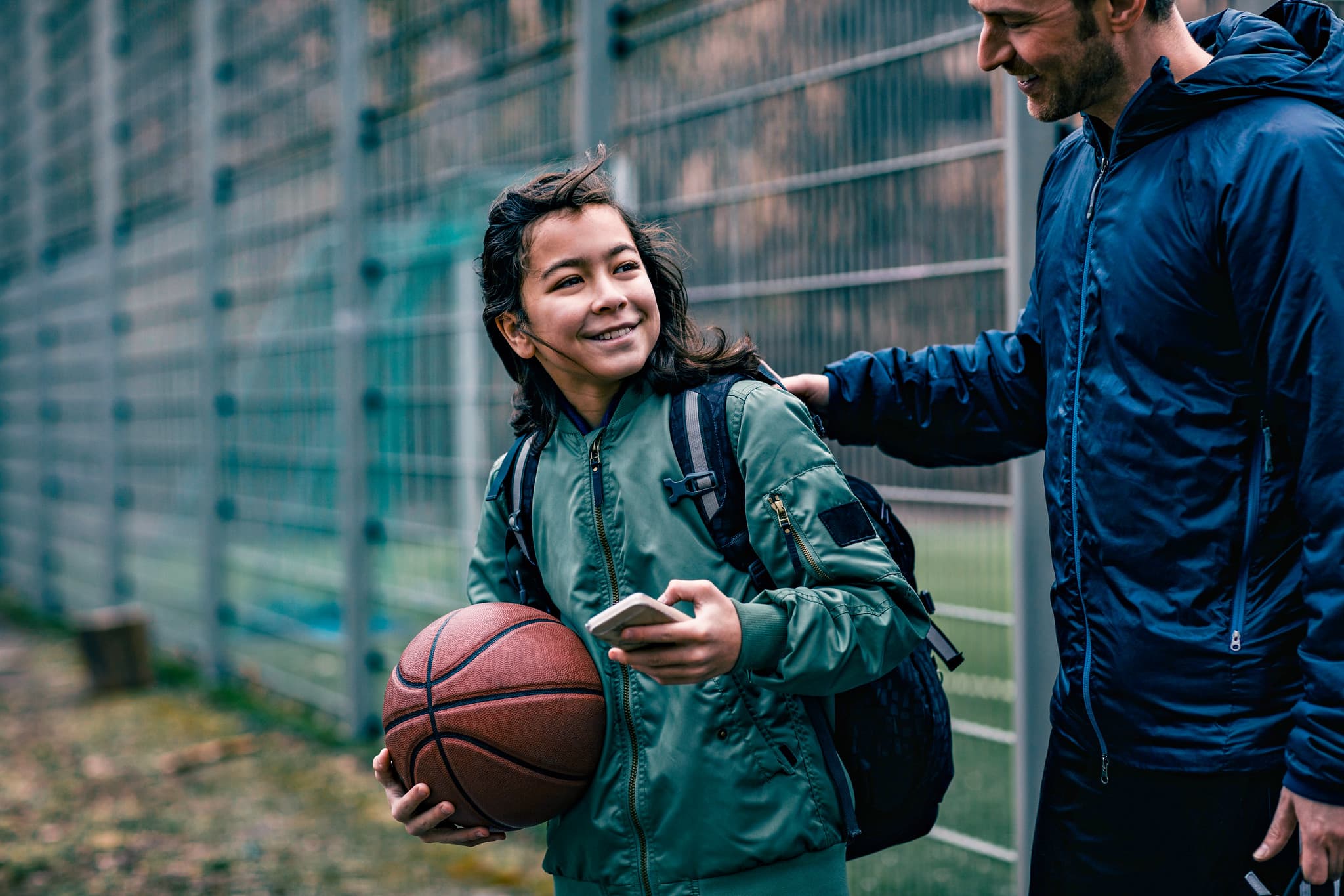 En jente i grønn jakke som bærer på en basketball. Hun smiler opp mot en voksen mann som har hånden sin på skulderen hennes