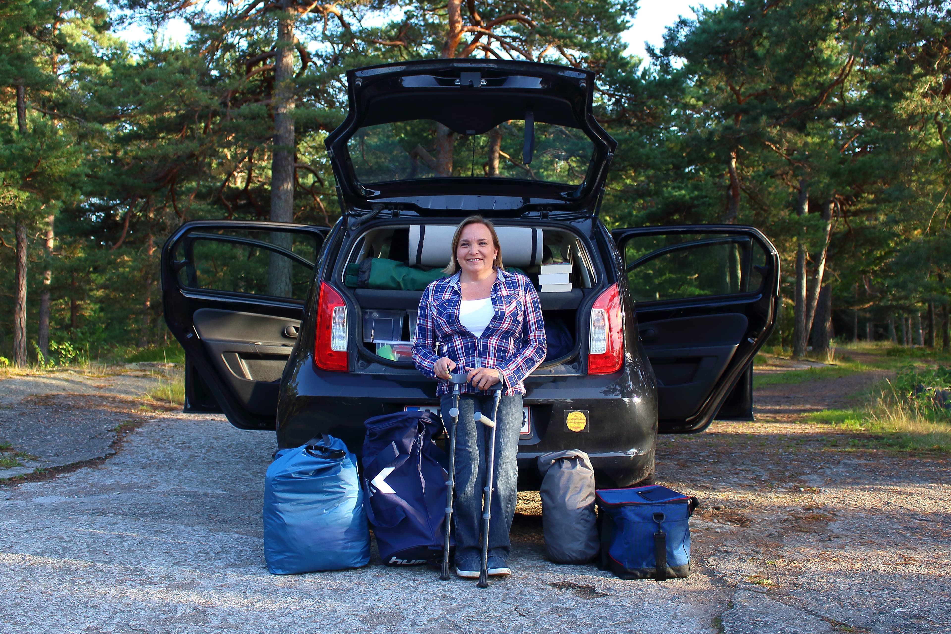 Adele Heidenreich, i rutete skjorte og med krykker, lener seg mot en bil som står på en skogsvei,med åpent bagasjerom, dørene på vid gap, og bagasje på bakken rundt seg.