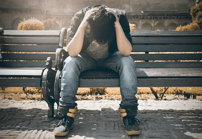 Ung person som sitter på benk med hodet i fanget og hendene på hodet