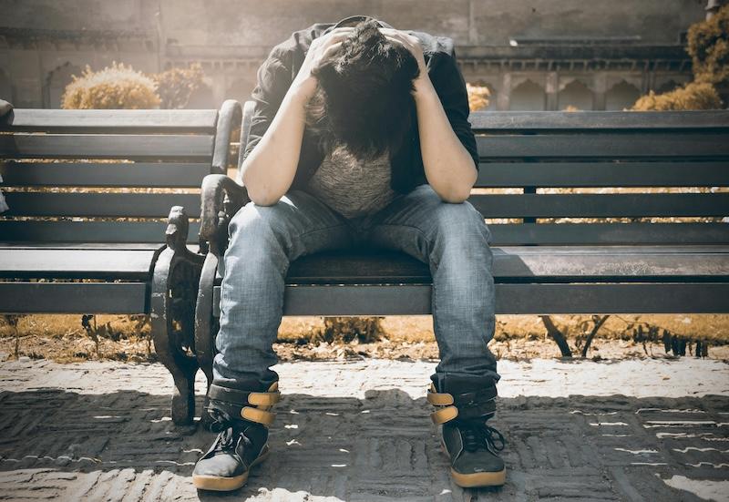 Ung person som sitter på benk med hodet i fanget og hendene på hodet