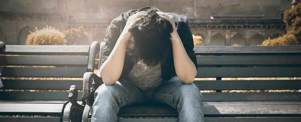 Ung person som sitter på benk med hodet i fanget og hendene på hodet