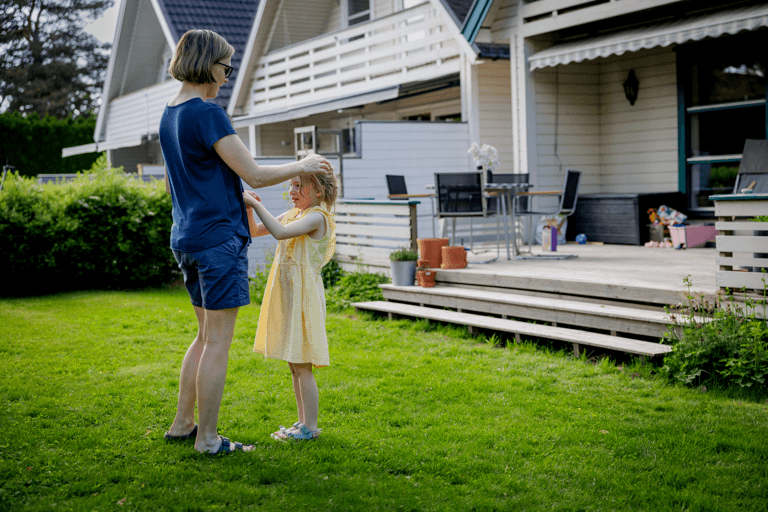 Bildet viser mor og barn stående på en plen foran et hus. De er vendt mot hverandre.
