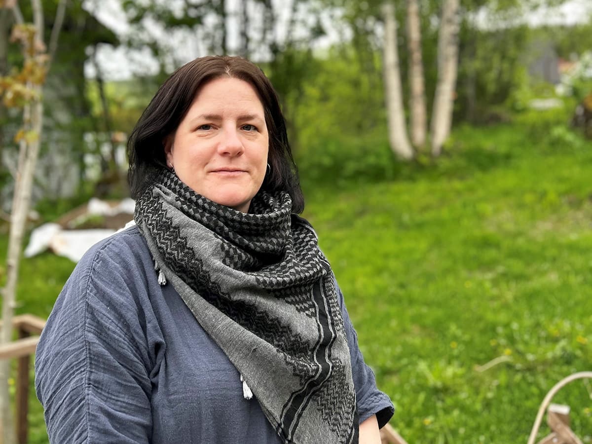 En mørkhåret kvinne i grå overdel og skjerf står foran en grønn plen