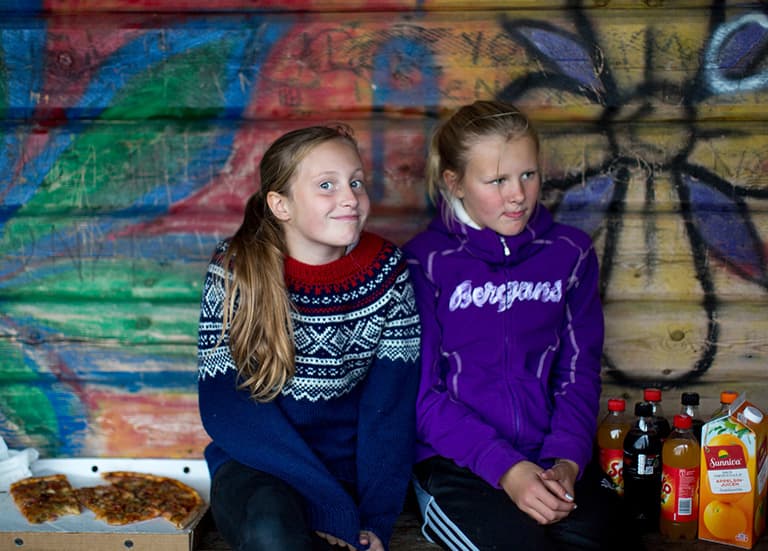 To ungdomsjenter med lyst hår som sitter på en benk. Ved siden av dem står det juice og pizza