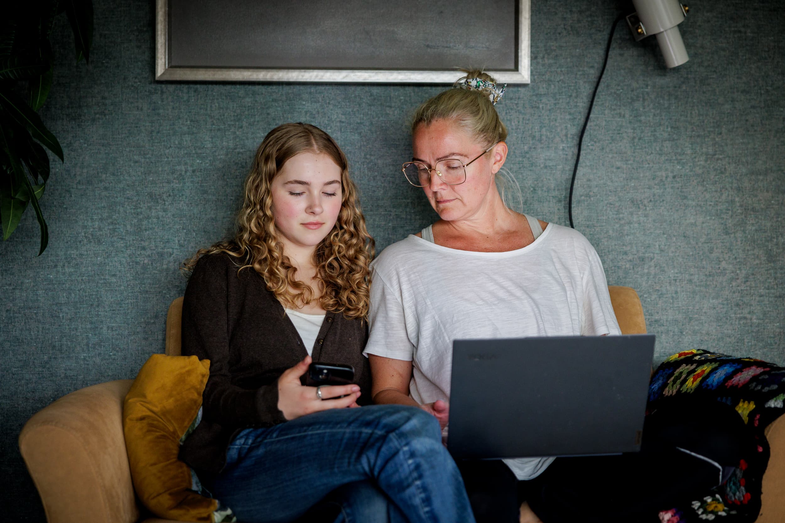 En ungdom sitter med en mobiltelefon mens en voksen ved siden av ser på skjermen, med en laptop i fanget