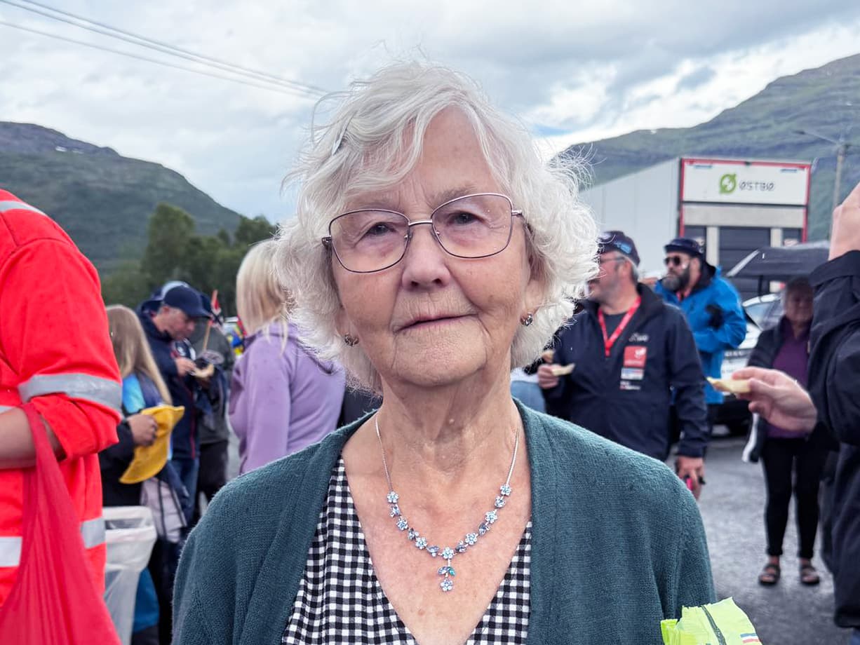 En eldre kvinne med hvitt hår og briller står foran mange mennesker