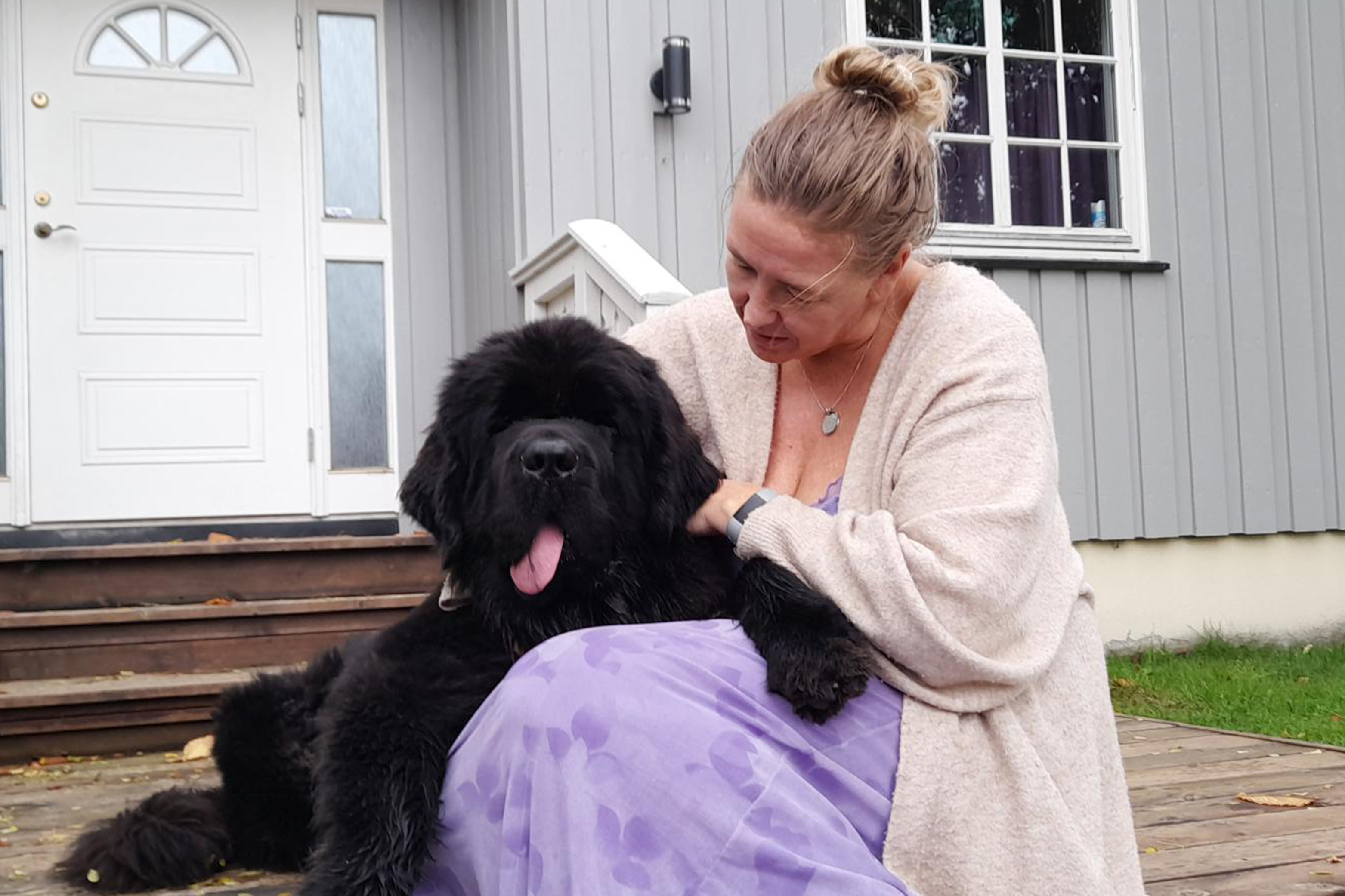En dame med lyst hår koser med hunden sin foran huset sitt