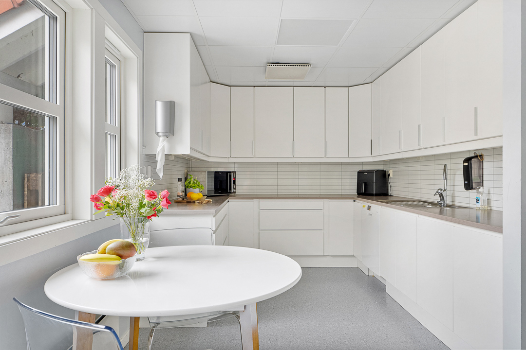 Kjøkkenkrok med hvite fronter, rundt spisebord med frukt og blomster. 