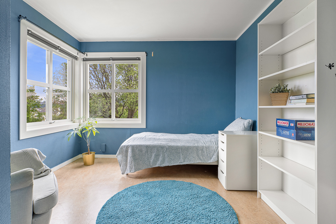 Ungdomsrom malt i blå farge, med send, bokhylle, nattbord, lenestol og plante, rundt teppe på gulvet. 